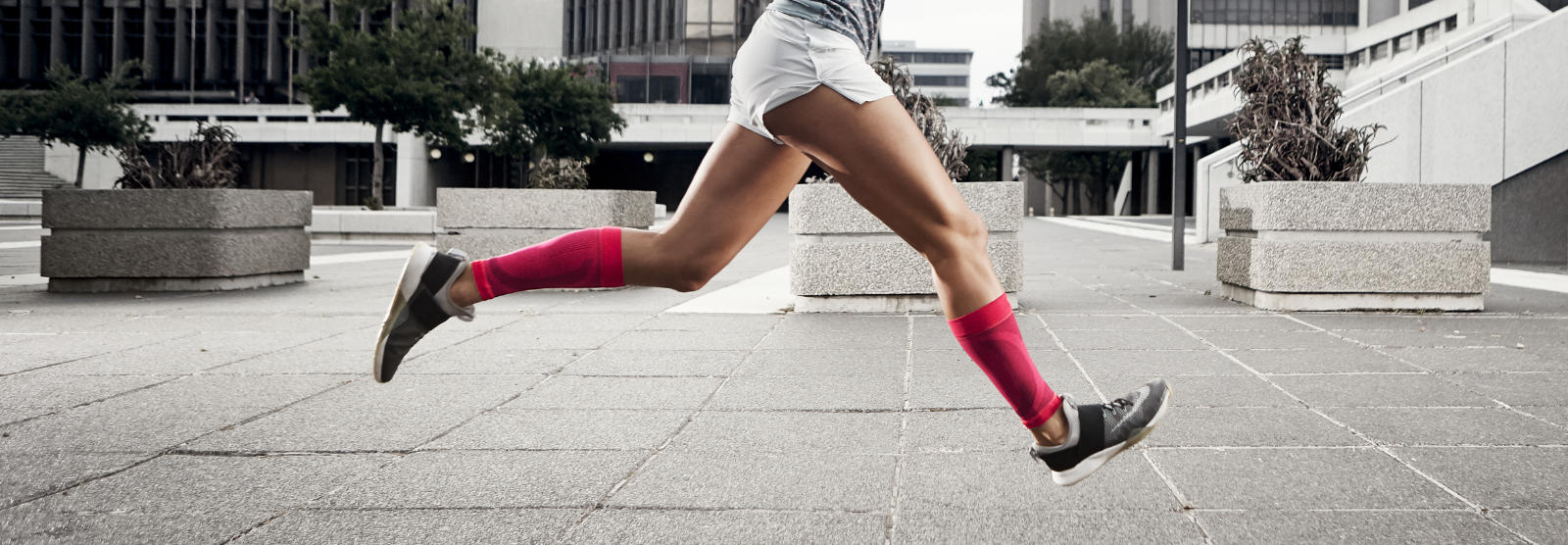 Briser la grisaille : nos manchons de compression en 7 couleurs Femme avec des manchons de mollet roses court rapidement avec les jambes en l'air à travers un désert urbain de béton