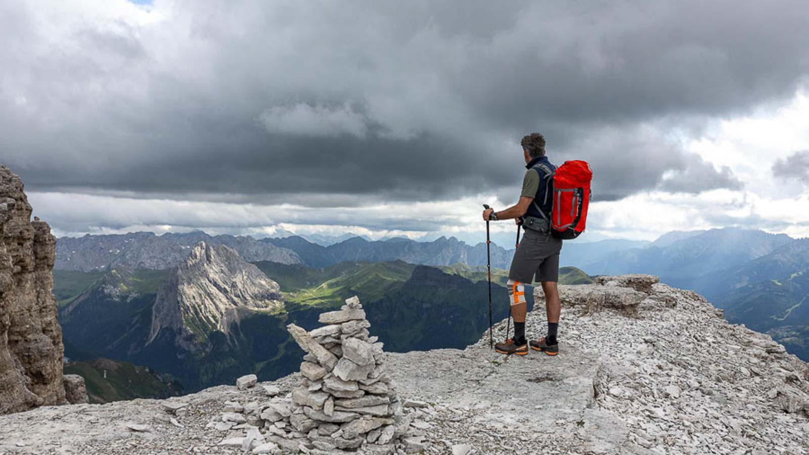 Randonneur sur un promontoire rocheux avec une genouillère de randonnée Une personne avec un sac à dos rouge se tient sur un promontoire rocheux dans les Dolomites et regarde au loin. Le ciel est nuageux et il y a une construction en cairn au premier plan.