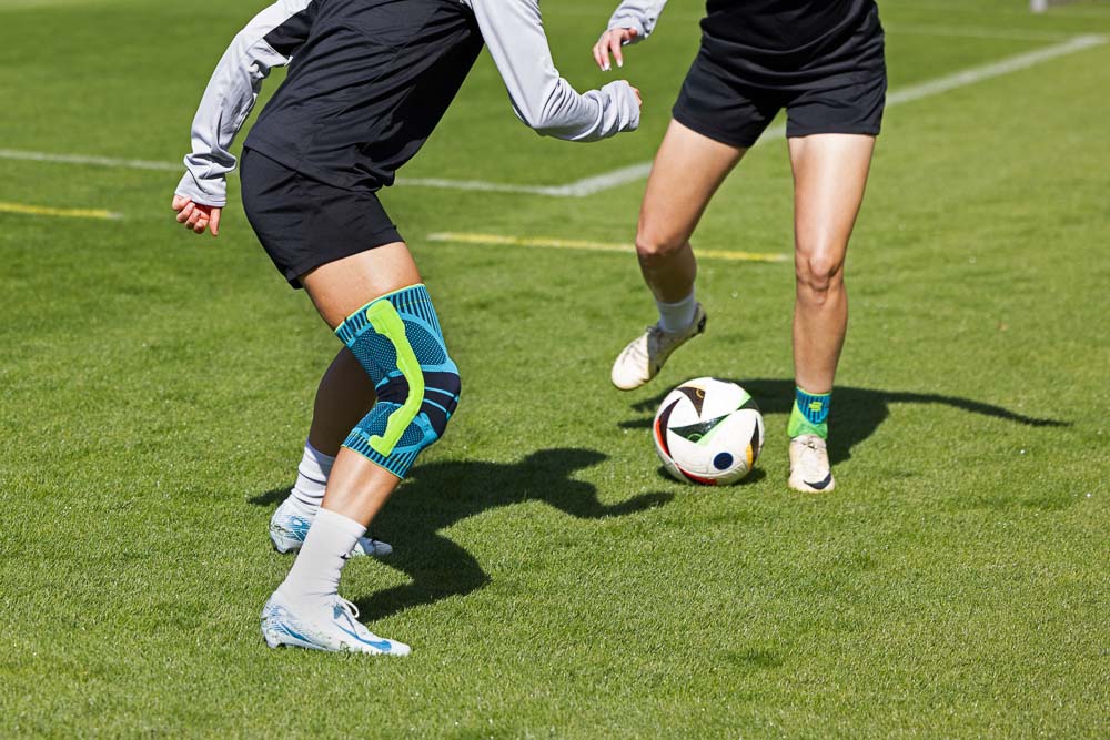 Zwei Fußball Spielerinnen sind im Zweikampf. Eine trägt eine Bauerfeind SPorts Knie Bandage, die andere eine Bauerfeind Sports Knöchel Bandage.