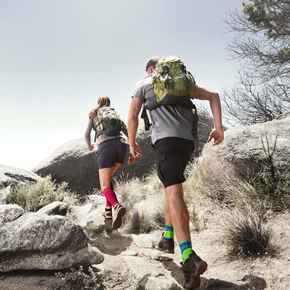 Hol Dir Unterstützung für Deine Wandertour: Zum Beispiel unsere Compression Sleeves und die Ankle Support Pärchen mit pinken Wadensleeves und blauen Sprunggelenkbandagen wandert im Sommer zwischen Felsen einen kleinen Anstieg nach oben