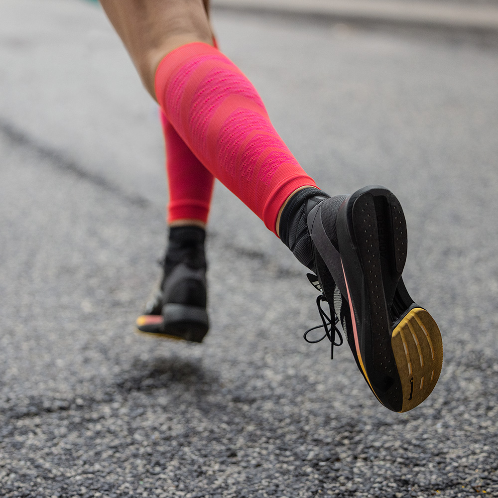Dynamischer Laufschritt mit roten Kompressions-Sleeves Detailaufnahme einer Läuferin auf Asphalt, die dynamisch nach vorne läuft. Sie trägt schwarze Laufschuhe und auffällige rote Run Performance Sleeves mit Kompression, die ihre Wadenmuskulatur unterstützen.