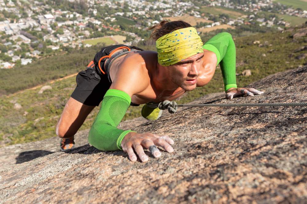 Klettern mit den Sports Compression Sleeves Arm Durch Seil gesicherter Kletterer am Berg	 freier Oberkörper	 Bandana am Kopf und grüne Arm Sleeves	 im Tal ist eine Stadt zu erkennen