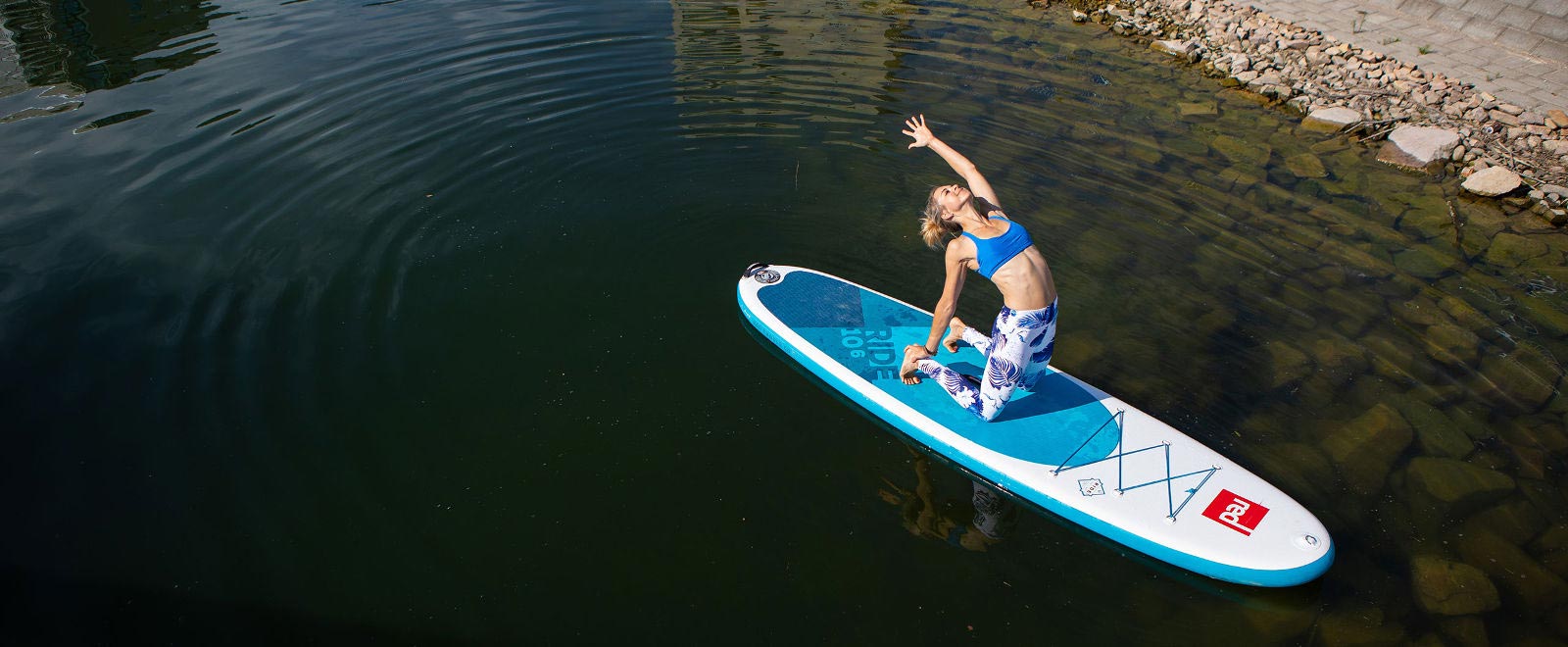 Une véritable tendance estivale : le SUP Yoga Une femme fait des exercices de yoga sur un SUP Board près de la rive, sous l'eau quelques pierres sont visibles