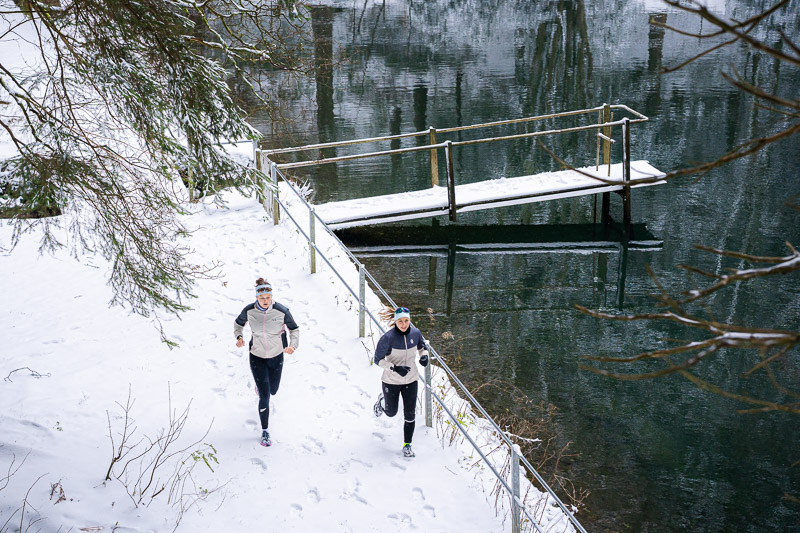 Winterlauf am See: Spuren im Schnee, Reflexionen im Wasser. Zwei Läuferinnen joggen entlang eines schneebedeckten Pfades neben einem See mit einem schneebedeckten Steg. Die Wasseroberfläche des Sees spiegelt die umliegenden Bäume wider. Beide Läuferinnen tragen helle Winterlaufjacken und schwarze Hosen.