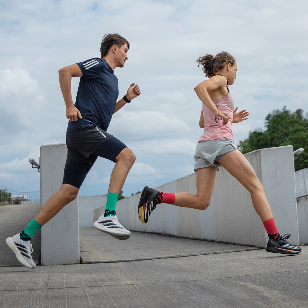Gemeinsam stark ins Ziel. Zwei Läufer in Bewegung auf einer Rampe, ein Mann in blauen Shorts mit grünen Bauerfeind-Laufsocken und eine Frau in grauen Shorts mit roten Bauerfeind-Laufsocken.