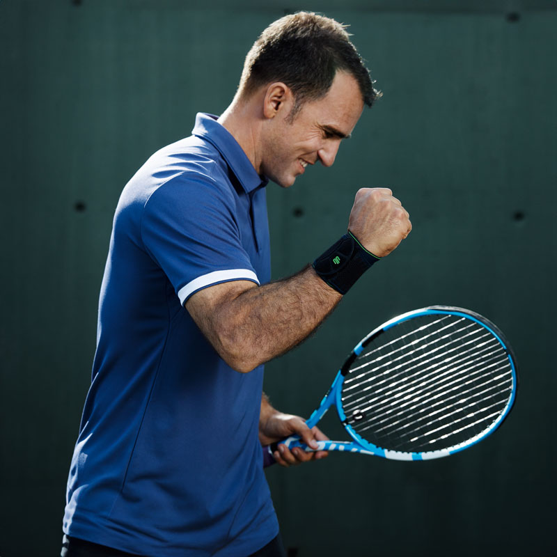 Mehr Stabilität und Präzision beim Tennis mit der Sports Wrist Support Tennisspieler mit schwarzer Handgelenkbandage hebt zufrieden seinen freien Arm und ballt die Faust