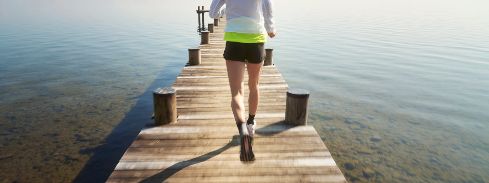 Schmerzen in der Kniekehle: Wo kommen sie her? Frau mit weißer Laufjacke und kurzen	 schwarzen Laufhosen läuft über einen Steg in Richtung Wasser