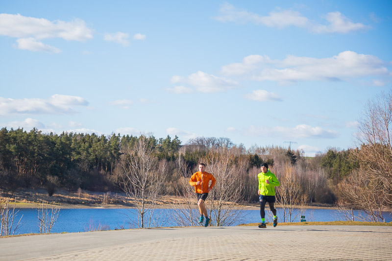 Frische Luft und freie Gedanken – Laufen durch die Natur Zwei Sportler laufen einen sonnigen Hügel hinauf, mit Blick auf einen ruhigen See und einen bewaldeten Horizont. Beide tragen leuchtende Laufjacken in Orange und Gelb.