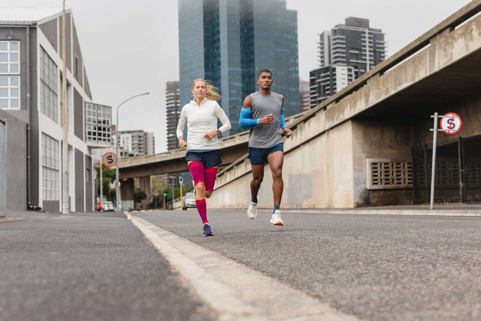 Que ce soit à Cape Town, Buenos Aires ou Tokyo : nos produits se font remarquer partout Une femme avec des compression sleeves roses et un homme avec des arm sleeves bleus et des chaussettes de course courtes courent à travers une grande ville