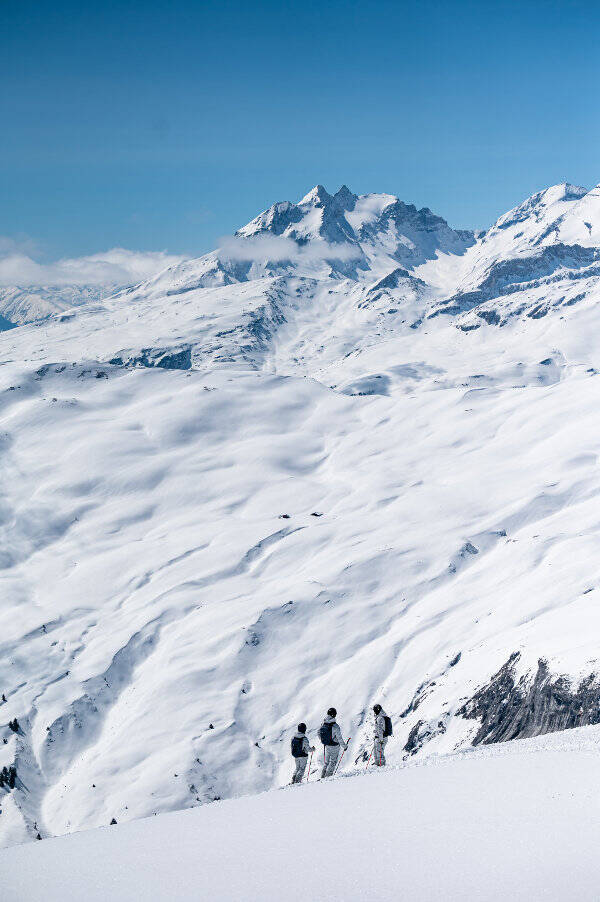 Les Alpes sont particulièrement majestueuses en hiver Panorama alpin enneigé	 devant lequel on distingue trois petits skieurs avec des sacs à dos