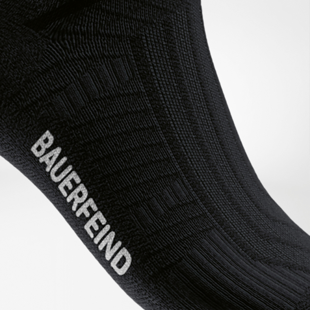 Mittelfußunterstützung und Rippstruktur bei der schwarzen Run Performance Socke Seitlicher Blick auf eine schwarze Sportsocke mit gut sichtbarer, integrierter Rippstruktur am Mittelfußbereich und weißem „BAUERFEIND“-Schriftzug – unterstützt das Fußgewölbe beim Laufen.