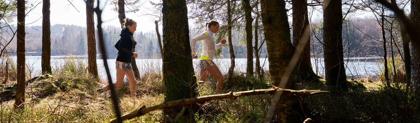 Knieschmerzen beim Joggen solltest Du nicht auf die leichte Schulter nehmen - auch wenn Deine Laufstrecke noch so verlockend ist Zwei Läuferinnen laufen hinter Bäumen und vor einem kleinen See hintereinander über einen Waldweg