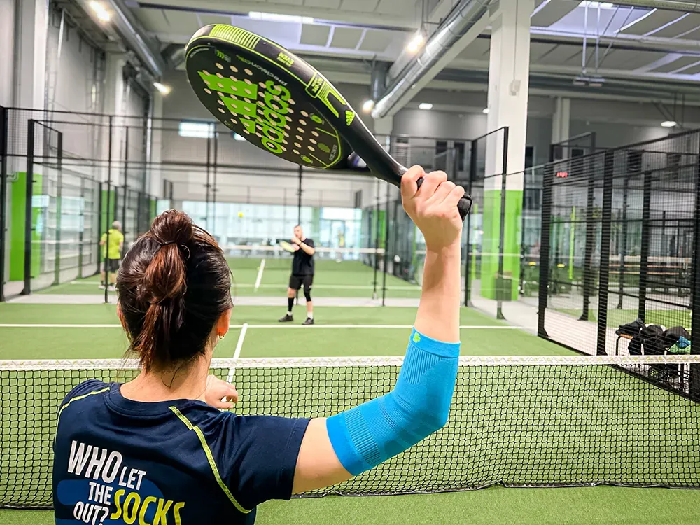 Mit Energie und Schutz am Netz! Eine Padel-Tennisspielerin steht am Netz und bereitet sich auf einen Schlag vor. Ihr Arm ist mit einer hellblauen Kompressionsbandage geschützt.