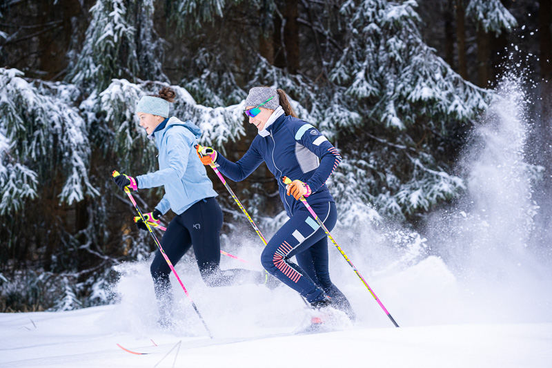 Dynamischer Skilanglauf im verschneiten Wald Zwei Frauen beim Skilanglauf durch verschneite Wälder. Sie tragen farbenfrohe Wintersportbekleidung und gleiten dynamisch durch den Schnee. Die Szenerie ist geprägt von dicht verschneiten Tannen, die das Winterwunderland unterstreichen.