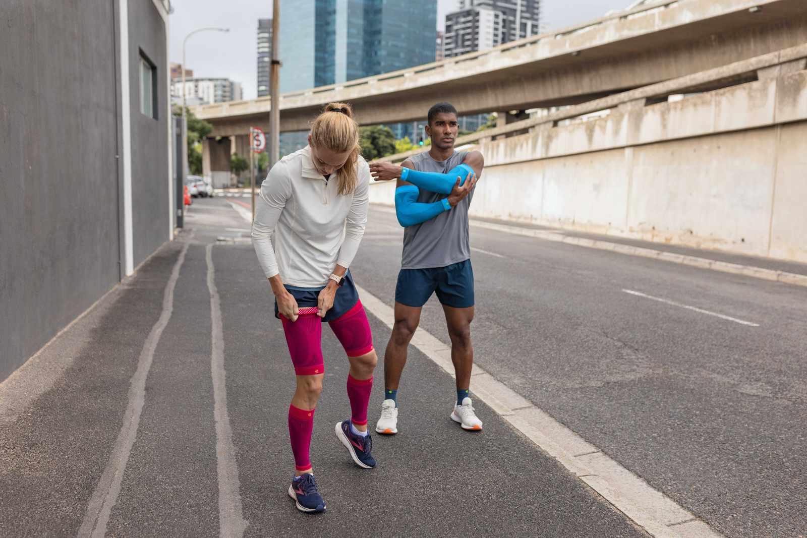 Même pendant la pause course, avoir du style : aucun problème avec nos produits stylés Une femme avec des Compression Sleeves roses et un homme avec des Arm Sleeves bleus et des chaussettes de course courtes prennent une pause de course au bord de la route dans une grande ville et s'étirent