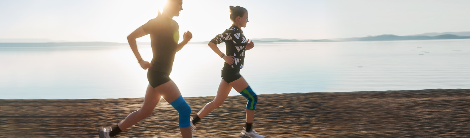 En cas de genou du coureur, une genouillère peut être un soulagement efficace Deux coureuses avec des genouillères courent au crépuscule sur une plage à côté d'un lac