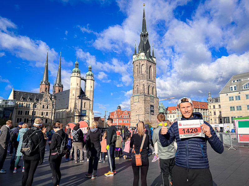 Ein Teilnehmer des Mitteldeutschen Halbmarathons steht auf dem Marktplatz in Halle und hält lächelnd seine Startnummer hoch. Als Zeichen seines Enthusiasmus zeigt der den Daumen nach oben. Er ist umgeben von Zuschauern und verschiedenen Bauwerken.