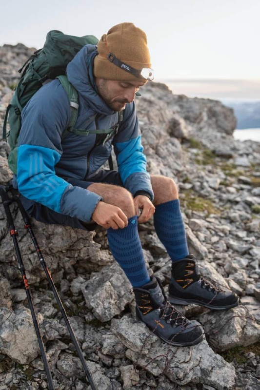 Gönn Dir die Schönheit der Natur und lass die Seele baumeln - mit unseren Produkten zum Wandern Lächelnde Wanderin mit Cappy und langen Wandersocken sitzt inmitten eines Feldes gelber Blumen