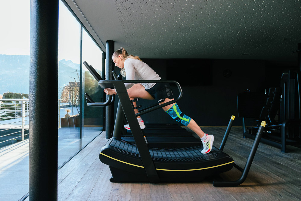 Sportlerin trainiert auf einem modernen Laufband in einem lichtdurchfluteten Fitnessraum. Sie trägt eine Kniebandage am rechten Bein, die das Knie während der intensiven Laufeinheit stabilisiert und unterstützt.