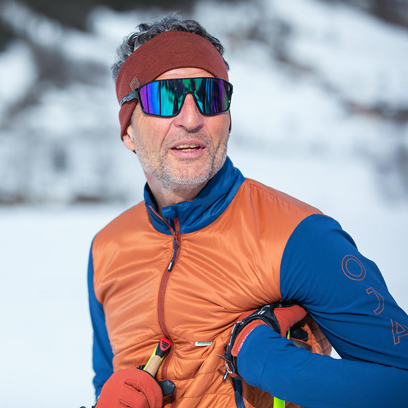 Porträt eines Skifahrers im Winterwald Ein älterer Mann, der eine Skibrille, ein braunes Stirnband und eine orange-blaue Winterjacke trägt, wurde in einem Portrait vor einem weißen Wald fotografiert.