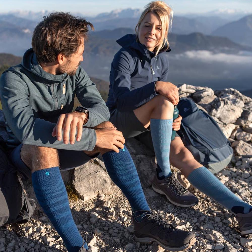 Höher hinaus mit Komfort – Entspannung auf jedem Gipfel. Zwei Wanderer genießen die Aussicht auf einem Berggipfel, während sie Merino Outdoor Kompressionssocken in Rot-Braun tragen. Die Socken bieten optimalen Komfort und Unterstützung für lange Touren in anspruchsvollem Gelände.