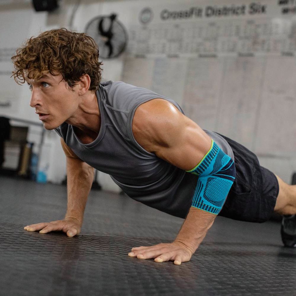 Stärke zeigen bei jeder Wiederholung! Ein sportlicher Mann mit muskulösem Oberkörper führt konzentriert Liegestütze in einem Fitnessstudio aus. Am rechten Arm trägt er eine blau-schwarze Ellenbogenbandage zur Stabilisierung und Unterstützung während des Trainings.