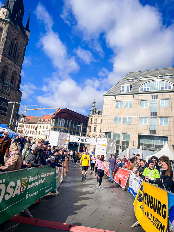 Die letzten Meter vor dem Ziel beim Halbmarathon in Halle, Zuschauer unterstützen die Läufer Die letzten Meter vor dem Ziel beim Halbmarathon in Halle. Läufer passieren das Ziel unter blauem Himmel, während das Publikum sie anfeuert.