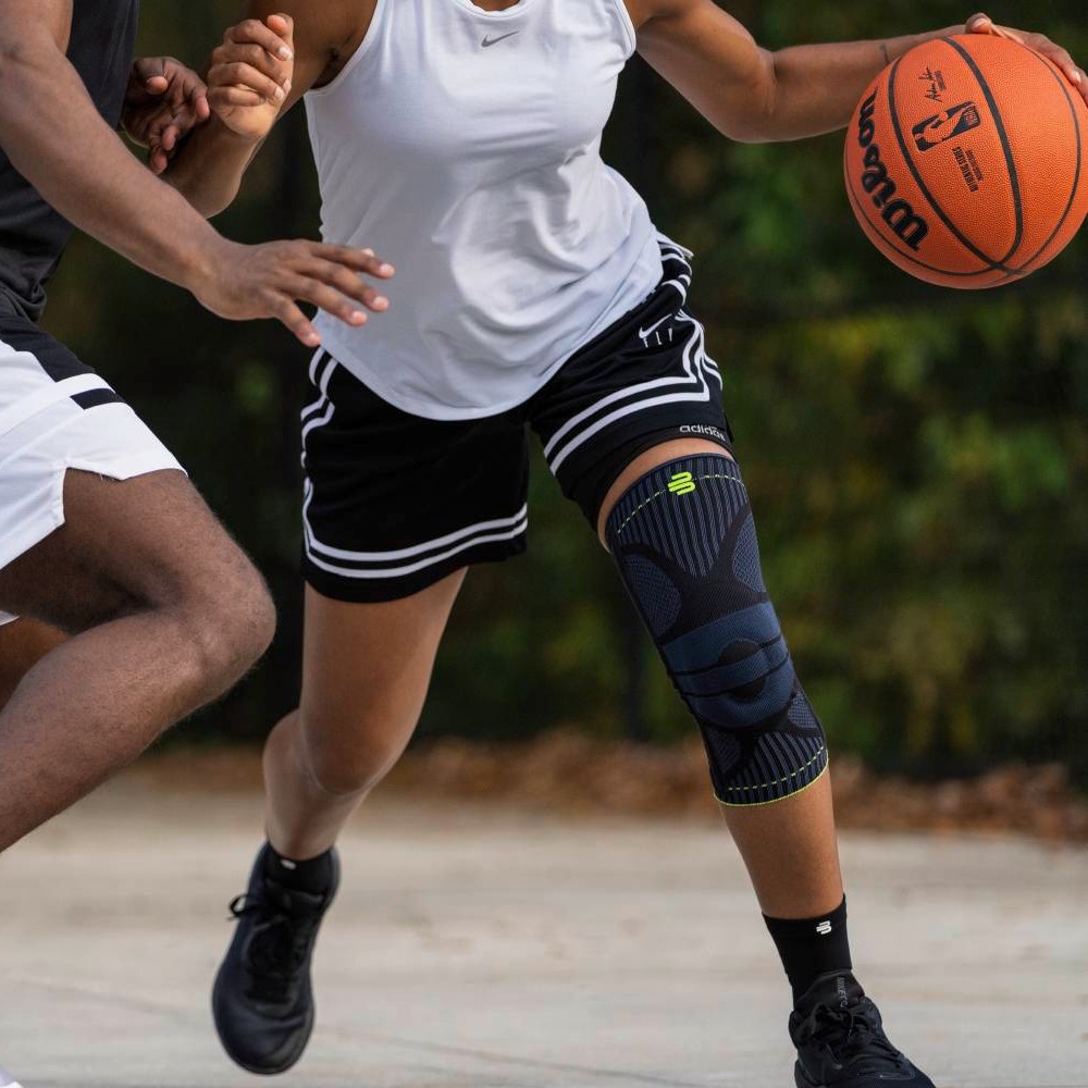 Schwarze Kniebandage im Einsatz auf dem Basketballplatz Eine Basketballspielerin trägt eine schwarze Kniebandage am rechten Bein, während sie in einer dynamischen Bewegung mit dem Ball in der Hand an einem Gegenspieler vorbeizieht.