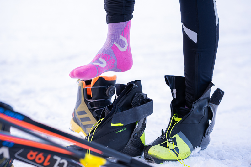 Perfekte Vorbereitung für den Langlauf mit passender Ausrüstung. Eine Detailaufnahme einer Skilanglauf-Ausrüstung: Eine Sportlerin zieht ihre neonpinkfarbenen Funktionssocken an, während neben ihr hochqualitative Langlaufschuhe und Skier auf dem Schnee liegen.