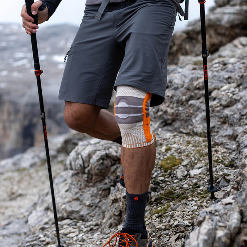 Wander Kniebandage im Einsatz Mann beim Tragen der Wander-Kniebandage in den Bergen beim Wandern mit Stöcken in felsigem Terrain.