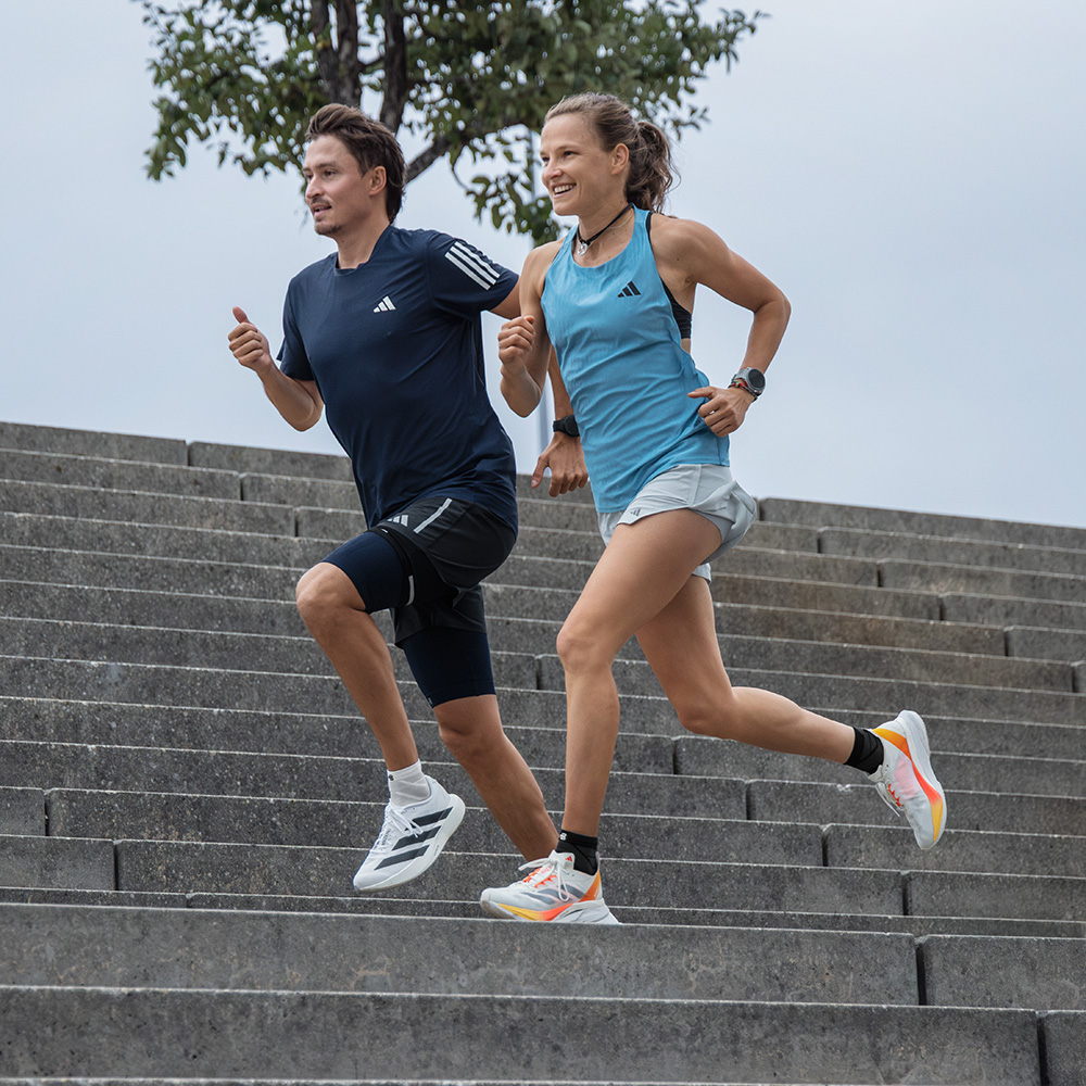 Mit Leichtigkeit die Stufen hinunter – gemeinsam in Bewegung Eine sportliche Frau und ein Mann laufen gemeinsam dynamisch eine breite Steintreppe hinunter. Beide tragen Sportkleidung und Schuhe von adidas.