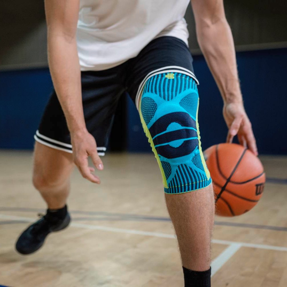 Basketballtraining mit türkisfarbener Kniebandage Ein Basketballspieler mit türkisfarbener Kniebandage auf dem rechten Bein dribbelt den Ball auf einem Indoor-Basketballplatz, der Fokus liegt auf dem Produkt und der Spielhaltung.