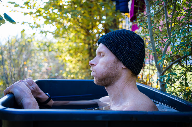 Mann beim Eisbaden in einem Outdoor-Bad. Ein Mann mit einer dunklen Strickmütze sitzt bis zu den Schultern in einem Eisbad im Freien. Er ist umgeben von herbstlicher Vegetation, und seine Gesichtsausdruck zeigt konzentrierte Atmung, um den Kältereiz besser zu verarbeiten.