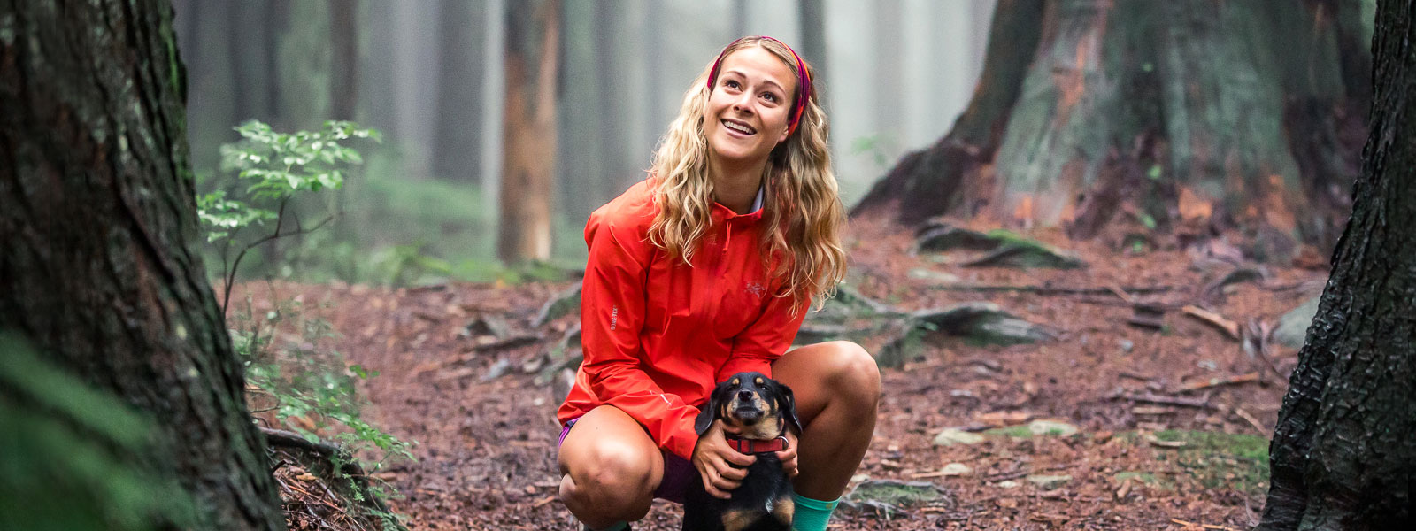 Idylle forestière avec chien : Hailey van Dyk en petite pause de course Femme avec une veste de course rouge et un bandeau tenant son petit chien sur un chemin forestier et souriant