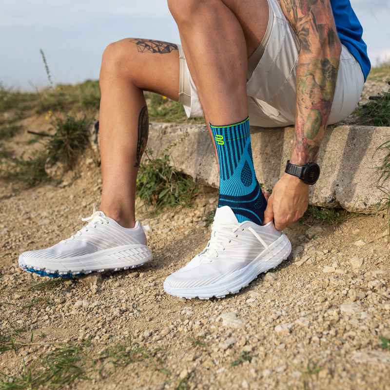 Mit Achillessehnenbandage noch sicherer und schmerzfrei laufen Mann zieht sich auf einem Stein sitzend einen Laufschuh über seine Achillessehnenbandage