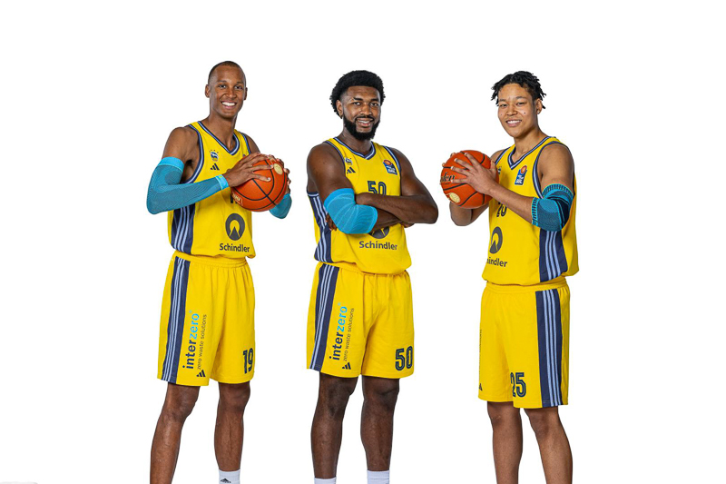 Alba Berlin Spielerteam – Offizielle Präsentation Drei Basketballspieler von Alba Berlin in ihren gelben Team-Trikots posieren mit Basketbällen vor einem neutralen weißen Hintergrund. Jeder Spieler lächelt und hält einen Basketball mit entspanntem, professionellem Ausdruck.