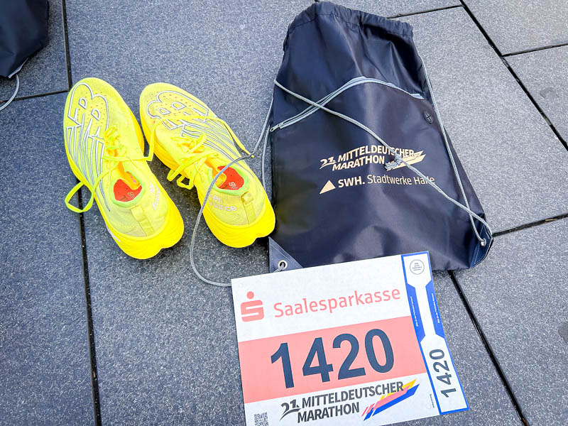 Halbmarathon-Ausrüstung - Startnummer und Laufschuhe am Renntag Renntag beim Halbmarathon: Gelbe Laufschuhe neben der Startnummer und einem Sportbeutel des 21. Mitteldeutschen Marathons, bereit für das Rennen.