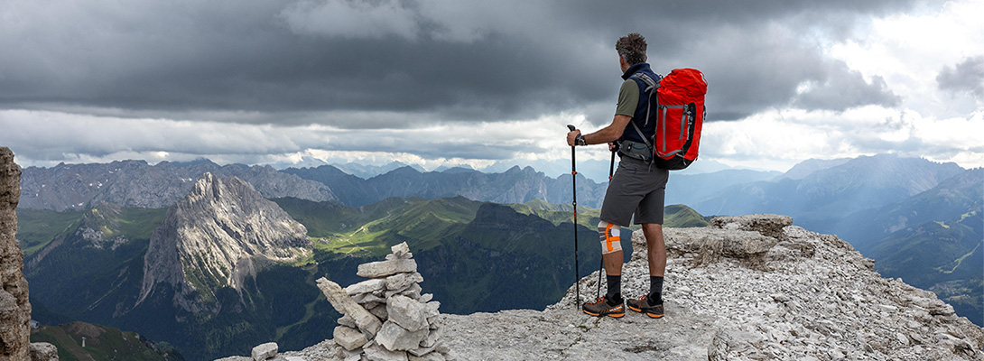 Mit der Wander Kniebandage ist jeder Gipfel stürmbar Bergsteiger mit rotem Rucksack und Wanderstöcken auf einer Bergspitze, er trägt eine Wander Kniebandage und blickt in die Ferne