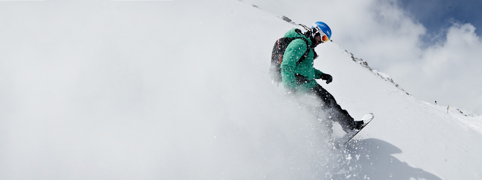 Snowboard-Equipment für noch mehr Schwung - hier bei uns Snowboarder mit grüner Jacke wirbelt beim Bremsen Schnee auf der die komplette linke Bildseite weiß färbt