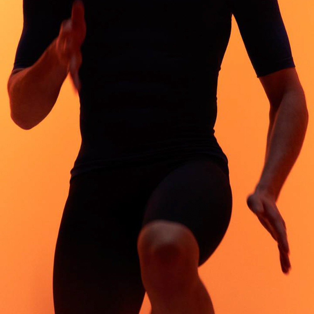 Fitnessübungen mit der neuen Bauerfeind Kompressionskleidung Ein Mann mit Sportbekleidung macht Kniehebelauf vor orangefarbenen Hintergrund
