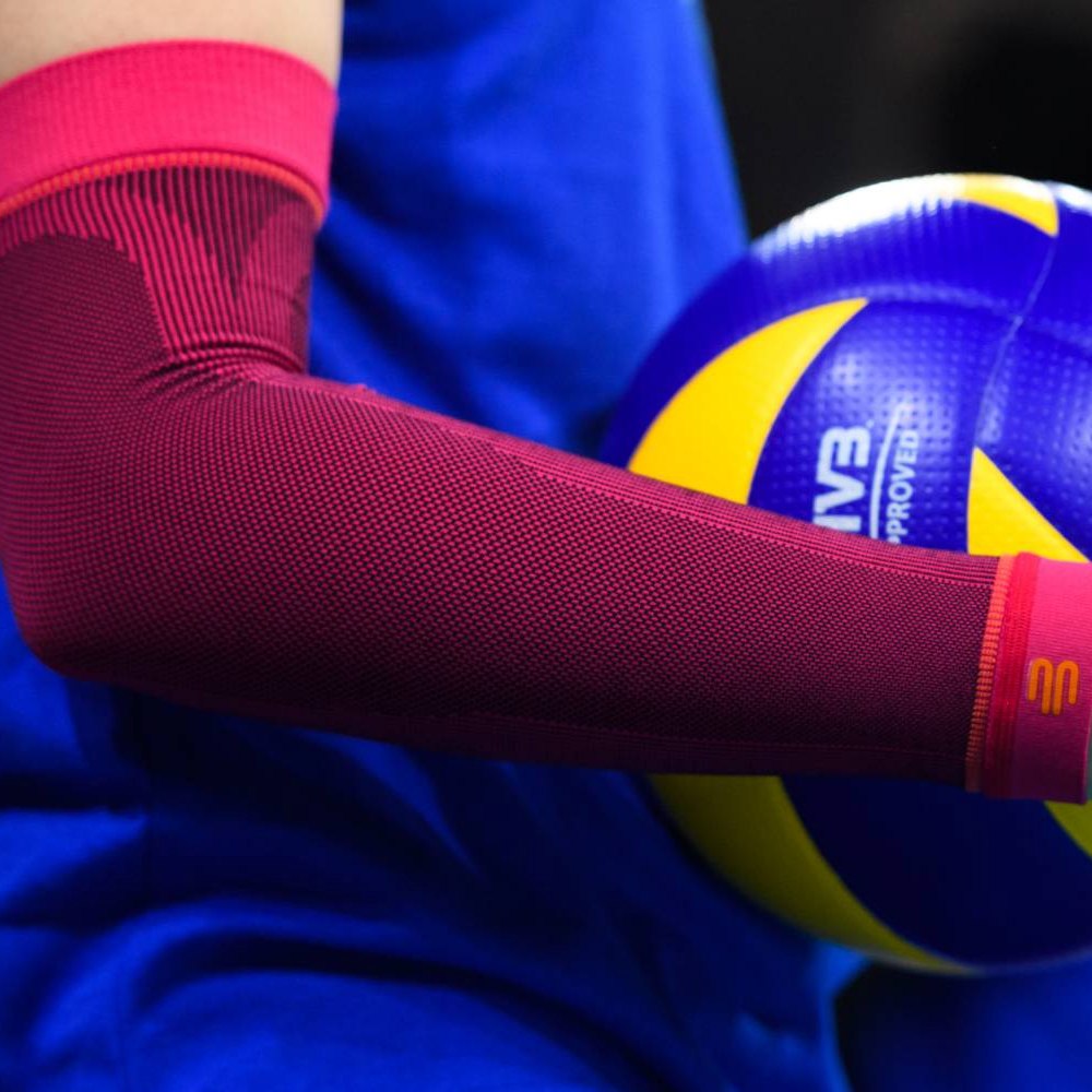 Volleyball Armsleeve in Pink beim Training Nahaufnahme eines pinkfarbenen Volleyball-Armsleeves an einem Spielerarm mit einem gelb-blauen Volleyball im Hintergrund.