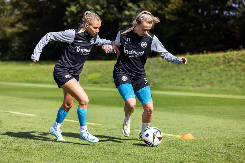 Zweikampf im Training bei Eintracht Frankfurt Zwei Spielerinnen von Eintracht Frankfurt kämpfen um den Ball während eines Trainingsspiels. Beide tragen schwarze Trainingskleidung mit Eintracht-Logo und blaue Kniebandagen von Bauerfeind Sports.