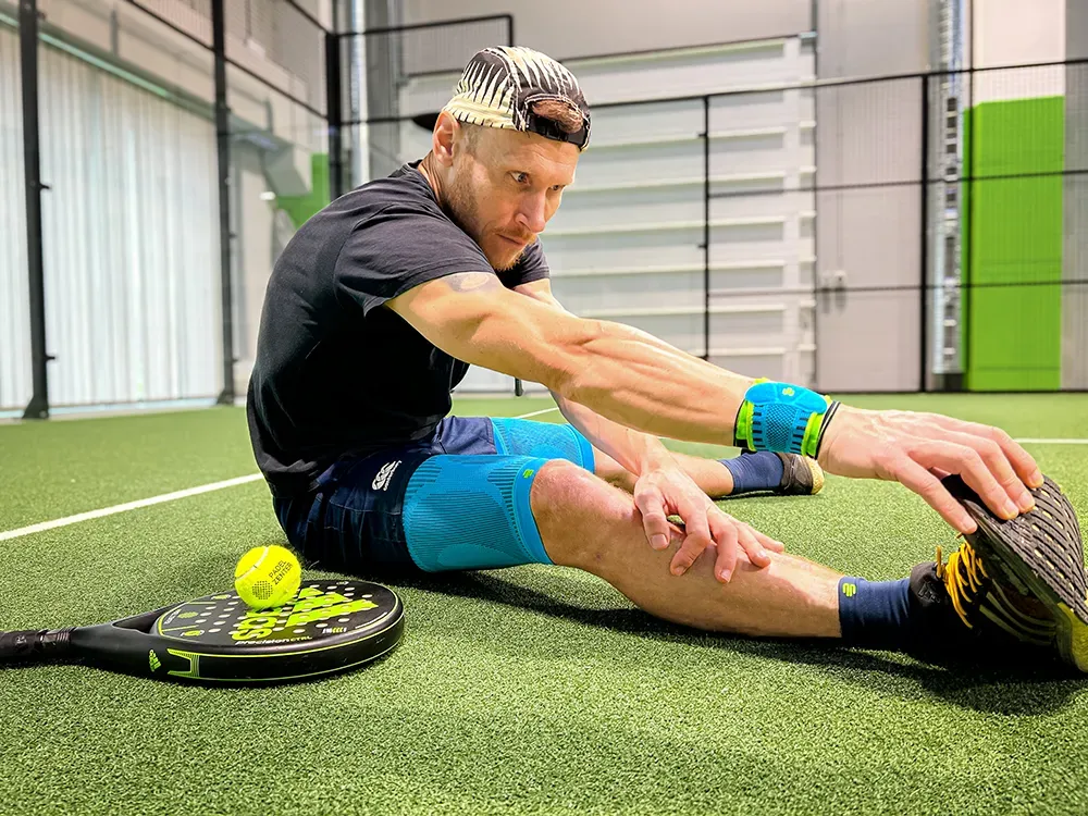 Fokussiert auf den nächsten Punkt – Stretching auf dem Padel-Court. Ein Sportler sitzt auf dem Boden eines Padel-Tennis-Platzes, dehnt seine Beinmuskulatur und trägt verschiedene Kompressionsprodukte an Arm, Bein und Handgelenk. Neben ihm liegen ein Padel-Schläger und ein Ball.