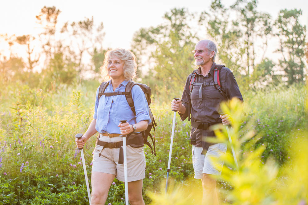 Aktives Wandern im Sommerlicht Ein älteres Paar wandert mit Trekkingstöcken durch eine blühende Sommerwiese im Abendlicht. Beide tragen leichte Wanderkleidung, Rucksäcke und sehen zufrieden aus.