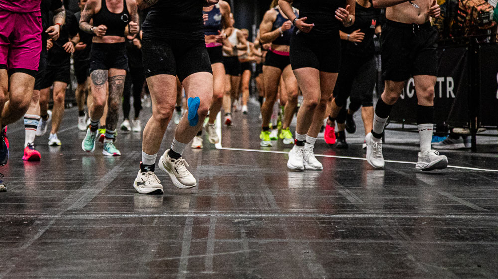 Dynamische Nahaufnahme der Beine mehrerer Athletinnen und Athleten, die auf einer dunklen Indoor-Laufstrecke im HYROX-Wettkampf laufen; Sportschuhe, Wettkampfkleidung und blaues Kinesiotape an einem Bein zeigen die intensive Bewegung