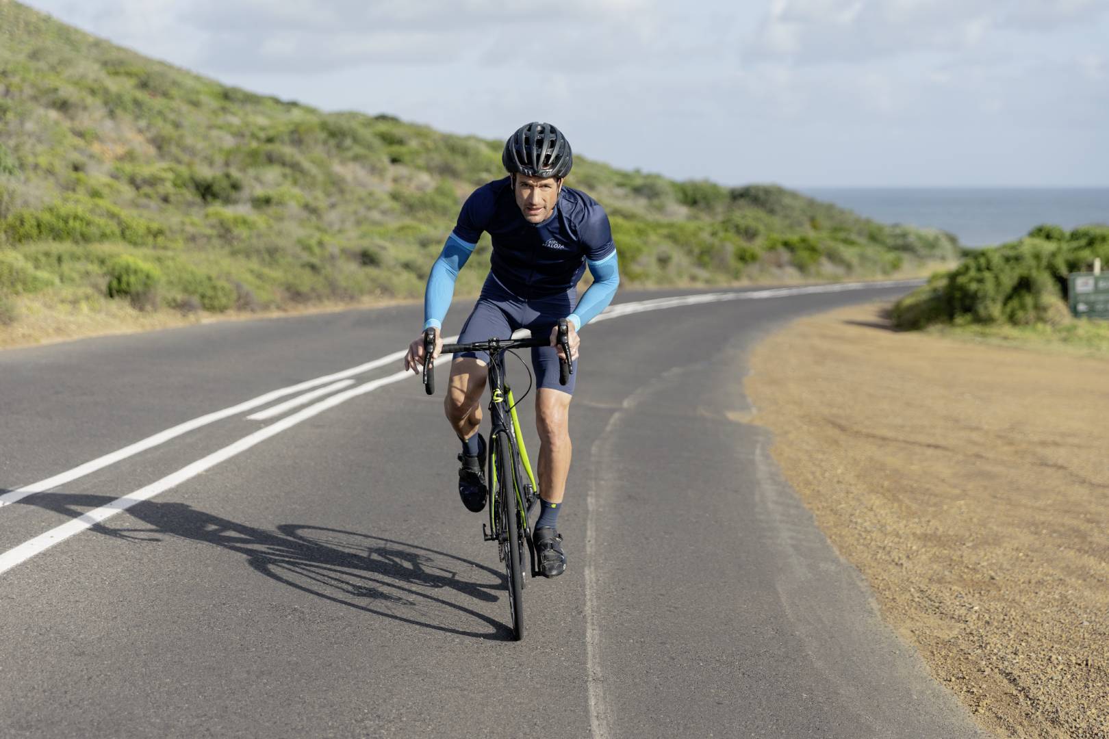 Un homme fait du vélo de course sportivement sur une route côtière par temps nuageux. Il est en danseuse.