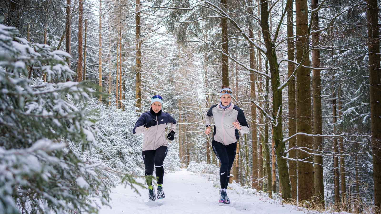 Winterlauf durch die verschneite Tannenlandschaft Zwei Personen joggen auf einem schneebedeckten Waldweg. Die verschneiten Tannen bilden einen dichten, winterlichen Hintergrund. Beide Läufer tragen warme Jacken und Stirnbänder für den Winterlauf.
