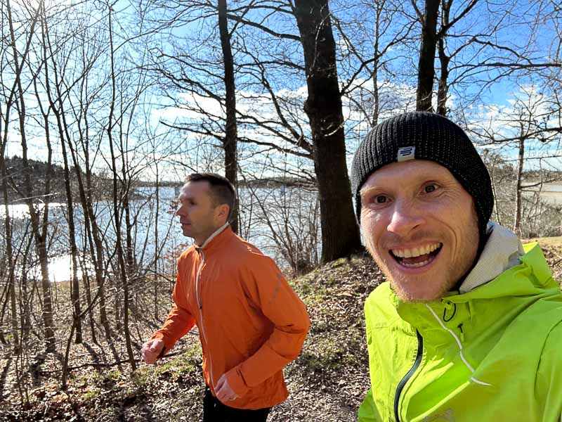 Laufend durch die Natur Zwei Männer laufen auf einem sonnigen Waldweg entlang eines Sees. Einer trägt eine neongrüne Jacke und eine Mütze, der andere eine orangefarbene Jacke.