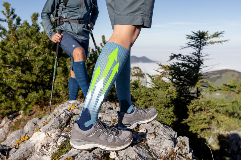 Zwei Wanderer auf einem felsigen Bergpfad mit Kompressionsstrümpfen, die ein dynamisches Design mit grünen und weißen Pfeilen zeigen. Die Strümpfe betonen die Unterstützung und den Energiefluss während der Aktivität.