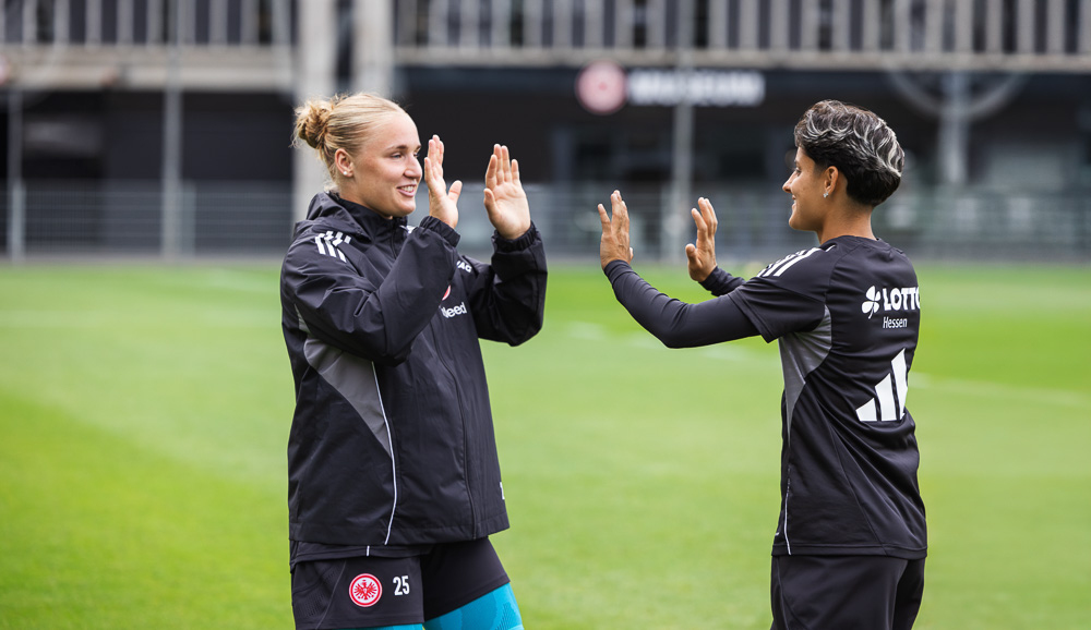 Zwei Fußballspielerinnen von Eintracht Frankfurt, gekleidet in Trainingskleidung, geben sich auf dem Spielfeld ein motiviertes High-Five. Beide tragen Kompressionsbandagen von Bauerfeind Sports.
